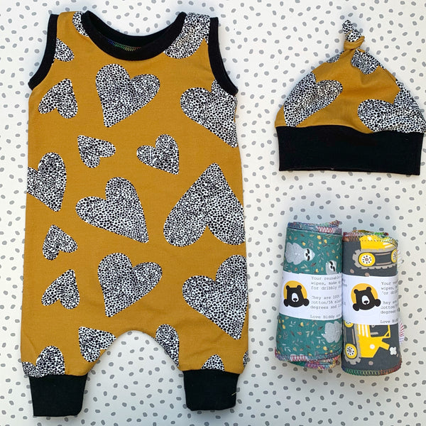 romper gift set contents - a romper and knotted hat in a mustard fabric with multidirectional hearts made of black dots on white. These items have a black trim. Plus two sets of 5 reusable wipes in mixed prints.