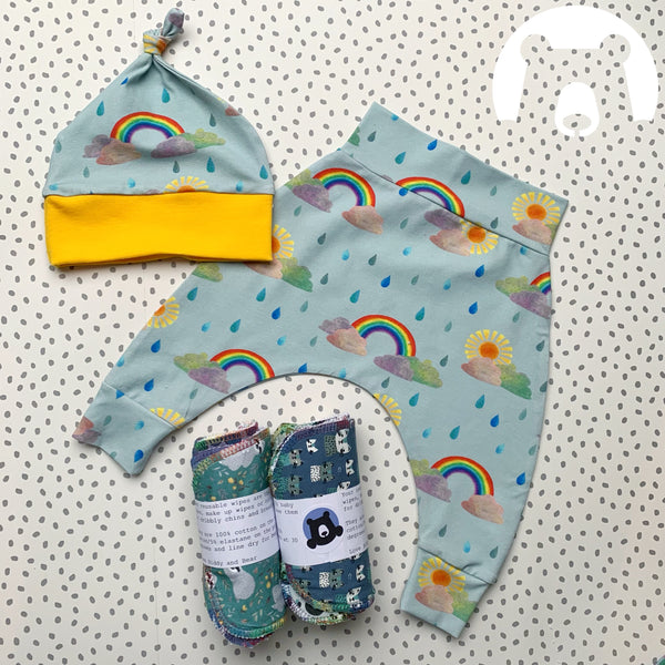 gift box contents - a knotted jersey hat, pair of harems and 2 sets of 5 reusable wipes in mixed prints. This set is made from a duck egg blue jersey with  watercolour rainbow, sunshine, cloud and raindrop elements.